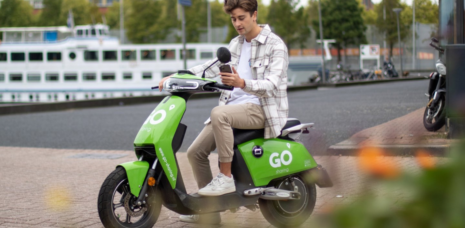 A man on a parked GO Sharing scooter. He is looking at his mobile phone.